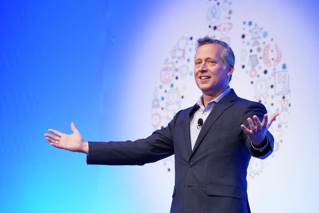 Motivational speaker engaging a corporate audience on stage with open arms and a bright blue, branded backdrop.