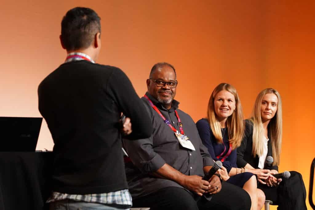 Panelists engage in a moderated corporate discussion on stage, captured with a warm backdrop. Captured by Christie’s Photographic Solutions.