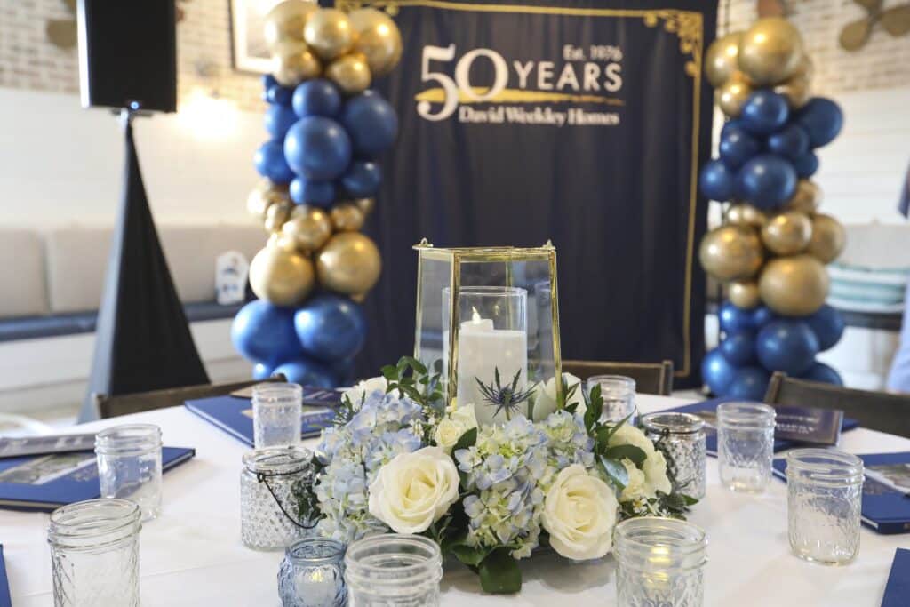 Elegant floral centerpiece with candle and blue and gold balloon backdrop celebrating a corporate anniversary, captured by Christie’s Photographic Solutions.