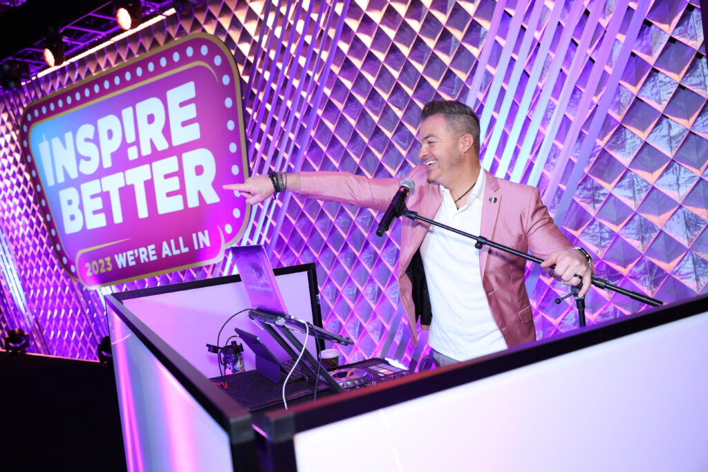 Corporate event DJ and emcee performing beside an illuminated Inspire Better backdrop with vibrant lighting, captured by Christie’s Photographic Solutions.