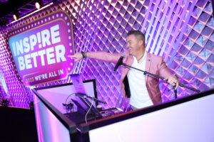 Corporate event DJ and emcee performing beside an illuminated Inspire Better backdrop with vibrant lighting, captured by Christie’s Photographic Solutions.