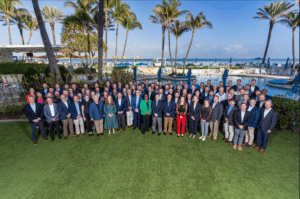 Large corporate group posed on a lawn beside an oceanfront resort pool with palm trees in the background, captured by Christie’s Photographic Solutions.