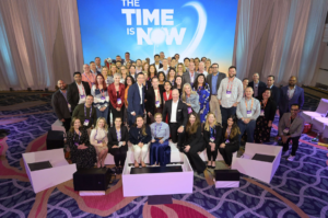 Large corporate attendee group portrait on a conference stage beneath a branded presentation screen, captured by Christie’s Photographic Solutions.
