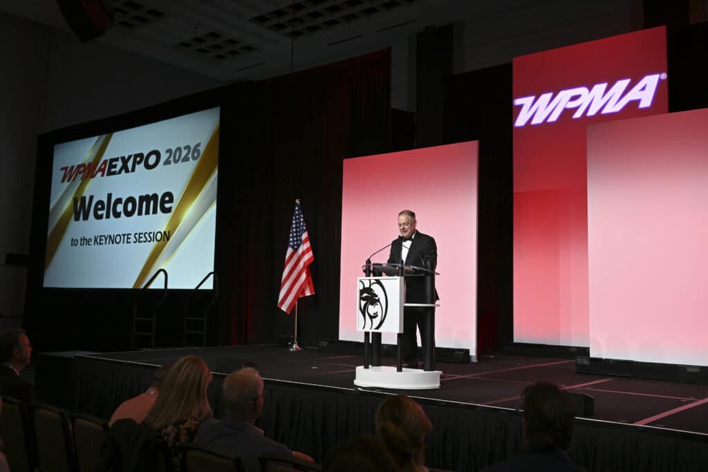 Executive speaker addressing attendees from the main stage during a keynote session with branded event screens, captured by Christie’s Photographic Solutions.