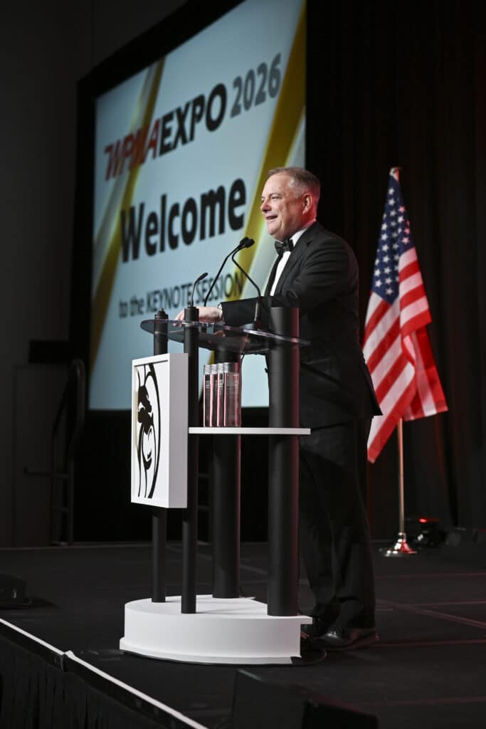 Executive speaker welcoming attendees from stage podium during a trade show opening session, captured by Christie’s Photographic Solutions.